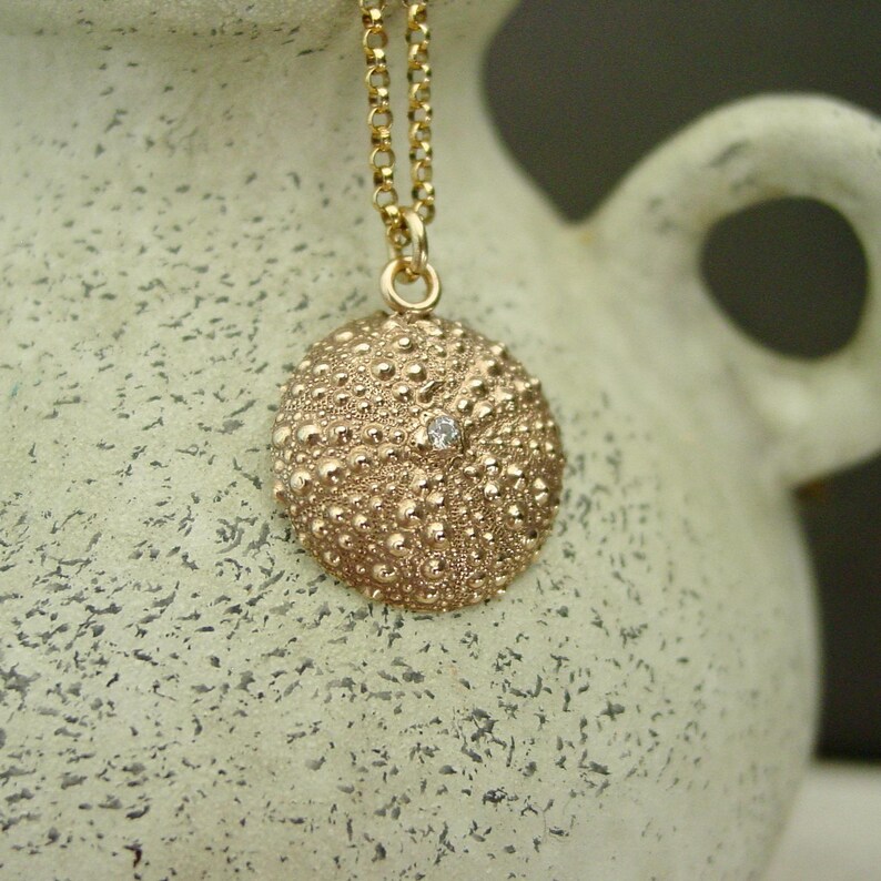 May include: A gold pendant necklace with a textured, round charm, reminiscent of a sea urchin. The charm has a small gemstone in the centre. The necklace chain is also gold, creating a coordinated and elegant design.
