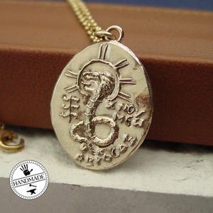 Może przedstawiać: Złoty owalny wisiorek na łańcuszku z wytłoczonym wzorem kobry i promienistymi liniami nad głową. Wisiorek zawiera wygrawerowane symbole oraz napis X NOV MEW i ABICAM, zawieszony na delikatnym złotym łańcuszku o drobnych ogniwach.