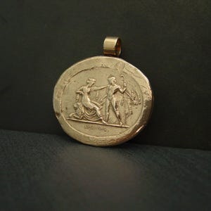 May include: Gold-colored pendant with a circular design featuring a relief of two figures in classical attire. One figure kneels, while the other stands with a staff. The pendant has a loop for a chain.