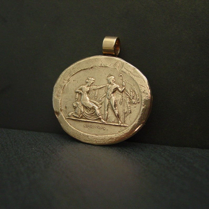 May include: Gold-colored pendant with a circular design featuring a relief of two figures in classical attire. One figure kneels, while the other stands with a staff. The pendant has a loop for a chain.