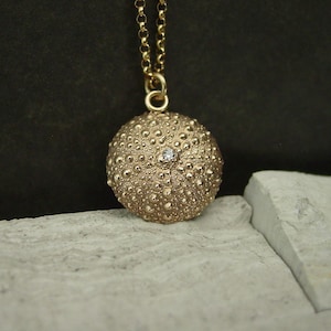 May include: A gold pendant necklace with a textured, spherical charm and a small gemstone. The charm hangs from a fine gold chain. The pendant has a detailed, bumpy surface. The necklace is gold in colour.