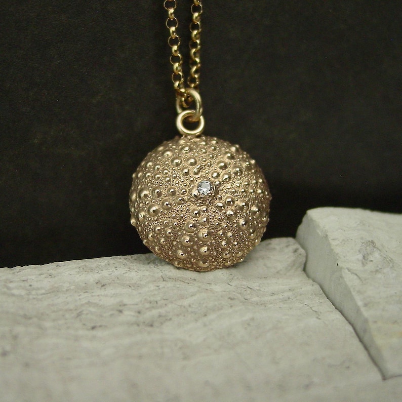 May include: A gold pendant necklace with a textured, spherical charm and a small gemstone. The charm hangs from a fine gold chain. The pendant has a detailed, bumpy surface. The necklace is gold in colour.
