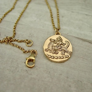 May include: A gold-toned necklace featuring a round pendant with a raised relief design of a mythical creature. The pendant hangs from a delicate rolo chain that includes a lobster claw clasp. The metal surface has a matte, textured finish.