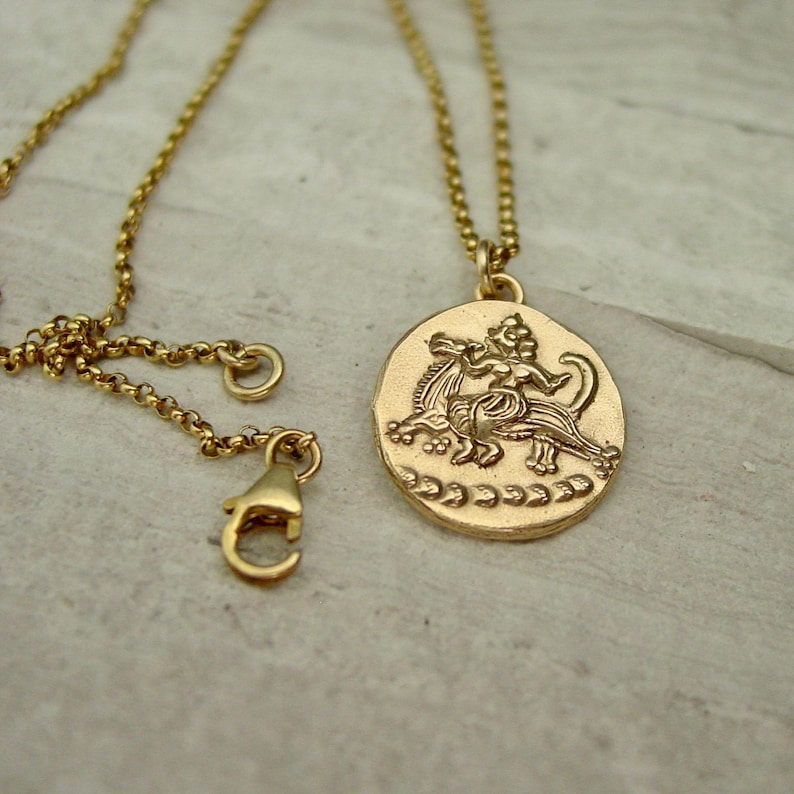 May include: A gold-toned necklace featuring a round pendant with a raised relief design of a mythical creature. The pendant hangs from a delicate rolo chain that includes a lobster claw clasp. The metal surface has a matte, textured finish.