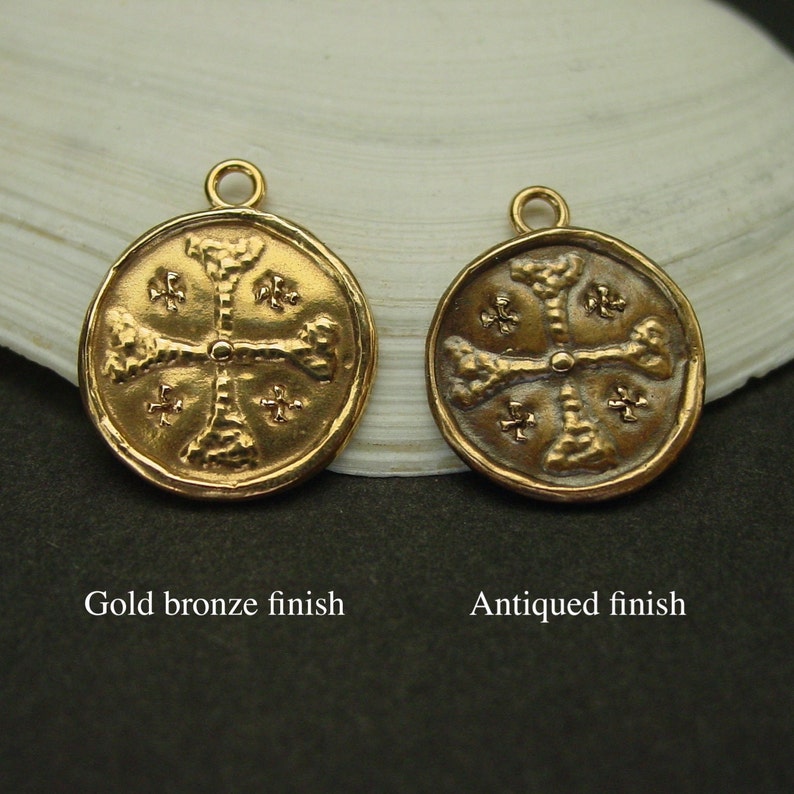 May include: Two round metal charms featuring a cross design with textured arms and small cross symbols in the quadrants. The left charm has a gold bronze finish, and the right charm has an antiqued finish. Each charm includes a small top loop.
