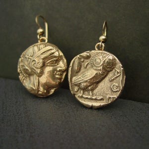 A pair of gold round coin earrings with cracks edges making it look ancient. On one coin is the raised image on an owl with a palm leaf and the moon with the letters AOE and on the other coin is the profile of a warrior woman wearing a helmet.