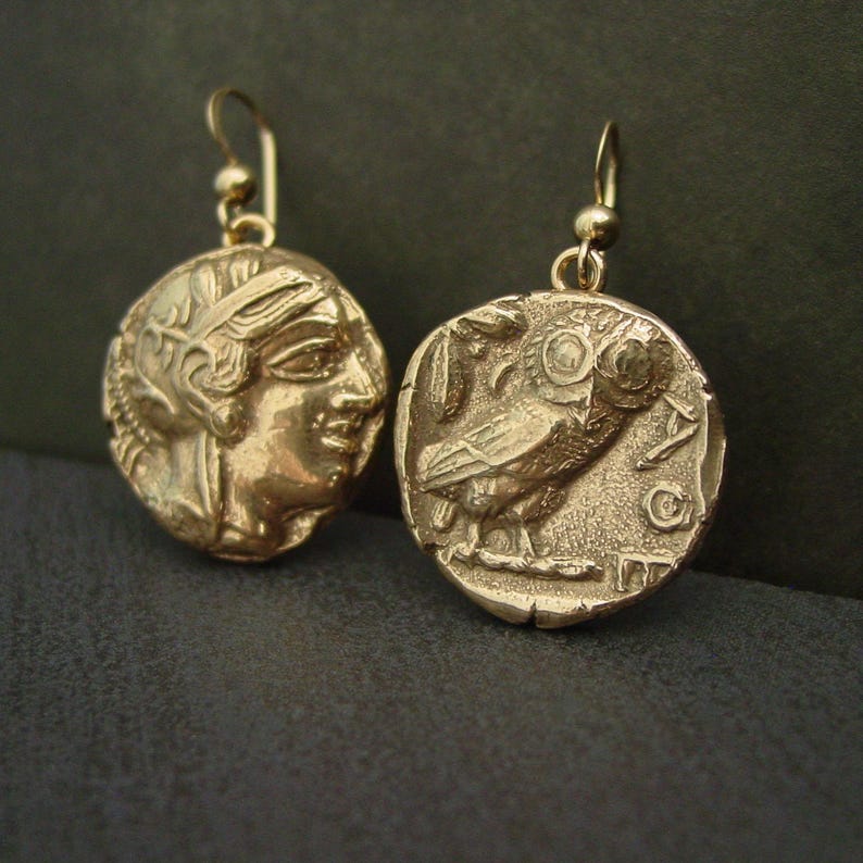 A pair of gold round coin earrings with cracks edges making it look ancient. On one coin is the raised image on an owl with a palm leaf and the moon with the letters AOE and on the other coin is the profile of a warrior woman wearing a helmet.