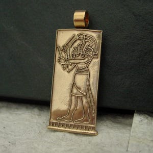 May include: A gold-colored rectangular pendant featuring an embossed image of an Egyptian deity. The deity is depicted in profile, holding objects, and wearing a headdress. The pendant has a loop at the top for a chain.
