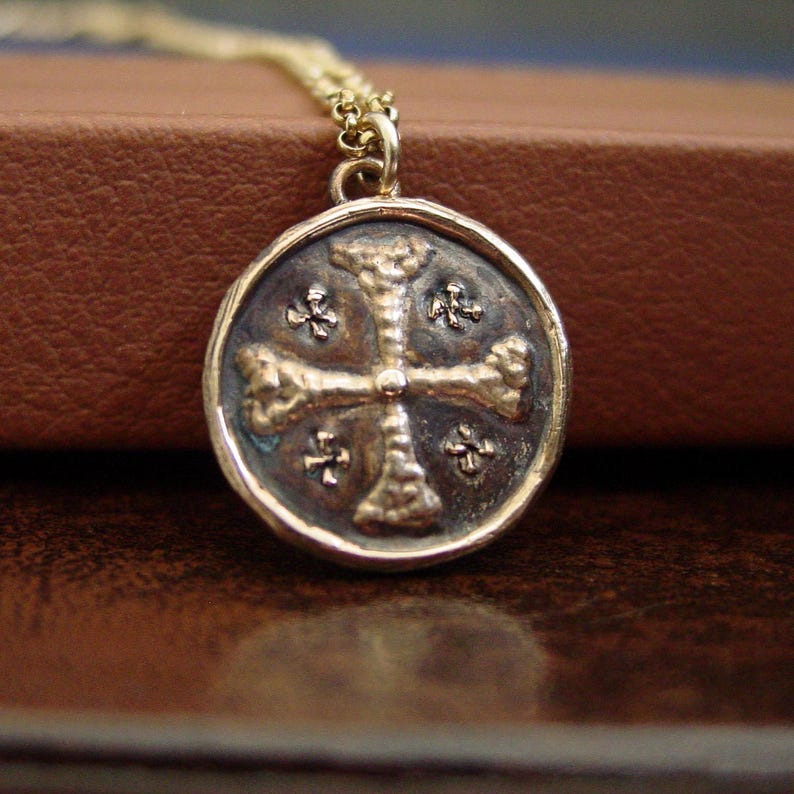 May include: A gold-toned necklace featuring a circular pendant with a textured, dark-toned centre. The pendant displays a raised, cross-shaped design with four smaller cross symbols positioned in the quadrants between the main cross arms.