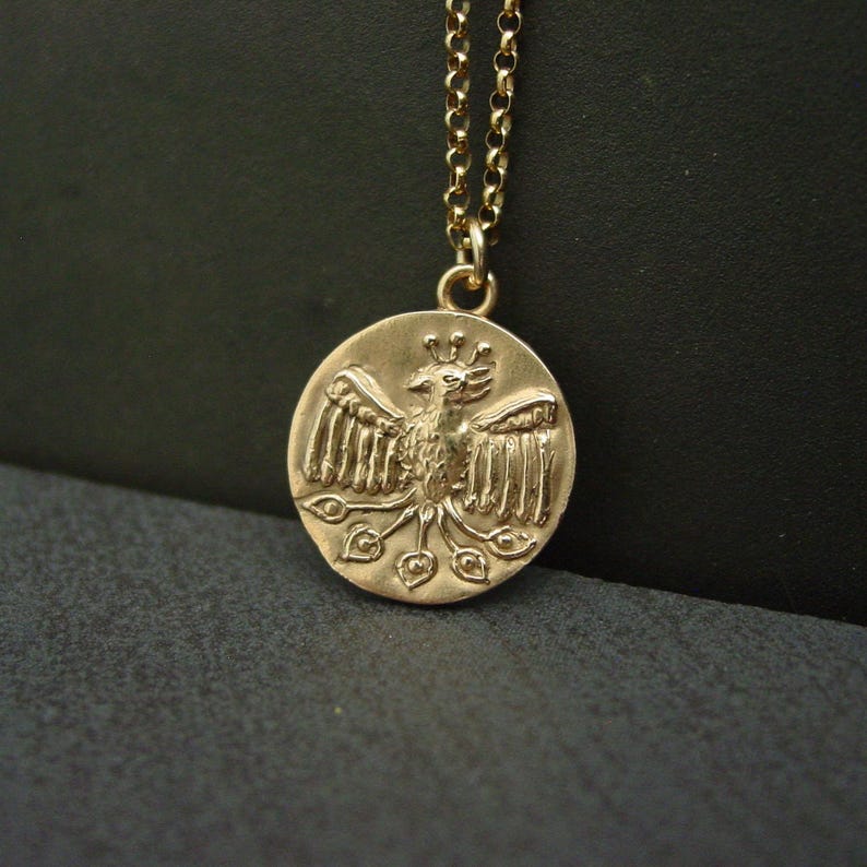 May include: A gold-toned circular pendant necklace featuring a relief design of a mythical bird with outstretched wings and a fanned tail. The pendant hangs from a delicate gold-toned rolo chain, showcasing a textured, metallic finish throughout.