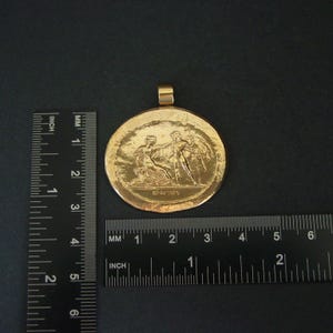 May include: A gold-colored pendant with a detailed relief scene of figures in classical attire. The pendant is round, approximately 2 inches in diameter, with a loop at the top for a chain. The background is dark.