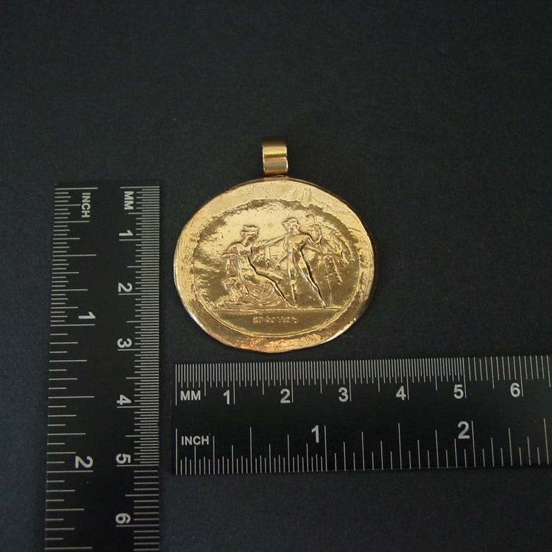May include: A gold-colored pendant with a detailed relief scene of figures in classical attire. The pendant is round, approximately 2 inches in diameter, with a loop at the top for a chain. The background is dark.