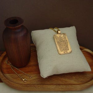 May include: A gold-toned rectangular pendant necklace featuring an engraved winged figure on the front. The pendant hangs from a delicate gold-toned chain, showcasing a detailed illustration of a standing character with large wings.