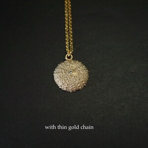 May include: A gold pendant necklace with a textured, round charm. The charm has a detailed, bumpy surface and a small, light-coloured stone. The necklace has a thin gold chain. The text "with thin gold chain" is at the bottom.
