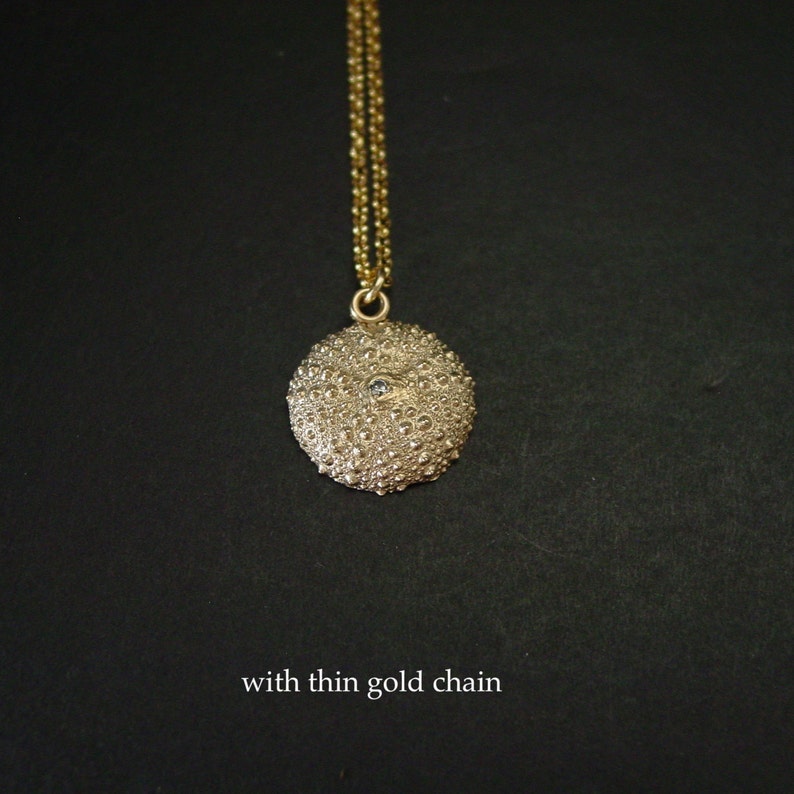 May include: A gold pendant necklace with a textured, round charm. The charm has a detailed, bumpy surface and a small, light-coloured stone. The necklace has a thin gold chain. The text "with thin gold chain" is at the bottom.