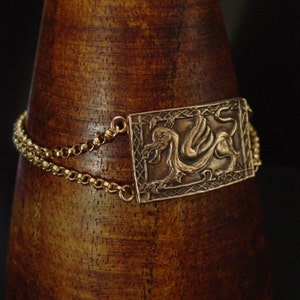 May include: A gold bracelet with a rectangular pendant featuring a detailed embossed image of a winged dragon. The pendant is surrounded by a decorative border.