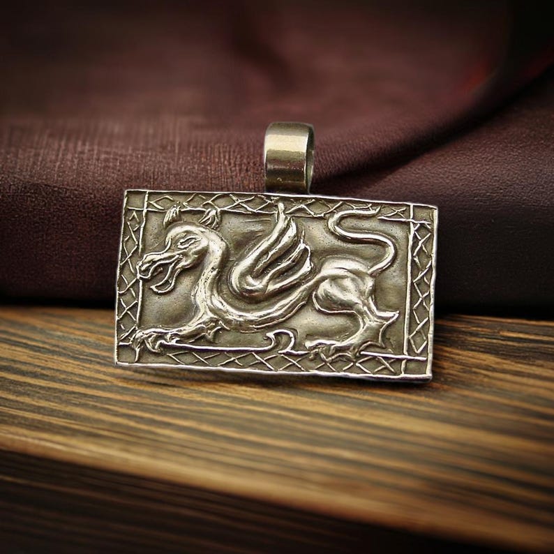 An antiqued rectangle bronze pendant with a wide loop on top for a chain to go through. On the pendant is an embossed Medieval style dragon crouching.