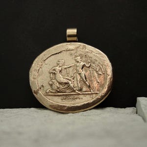 A large gold horizontal oval cameo pendant with a wide loop on top for a chain. On the pendant is a raised scene of a man holding a beast skin and a staff in front of a crowned woman seated on her throne with her lion behind her.