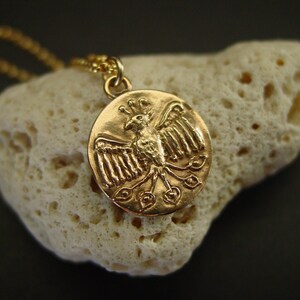 May include: A gold-toned circular pendant necklace featuring a relief design of a bird with outstretched wings and a fanned tail. The pendant is suspended from a matching gold-toned chain, showcasing a detailed metallic finish on the bird motif.