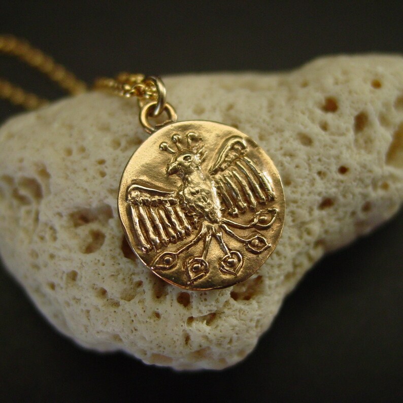 May include: A gold-toned circular pendant necklace featuring a relief design of a bird with outstretched wings and a fanned tail. The pendant is suspended from a matching gold-toned chain, showcasing a detailed metallic finish on the bird motif.