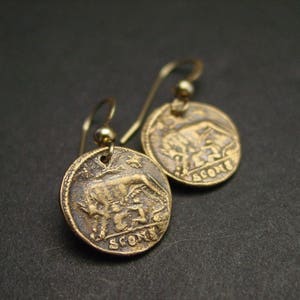 May include: Gold tone earrings featuring a round coin design with a raised image of a bull and a person. The coin has the word "SCONS" inscribed on it.