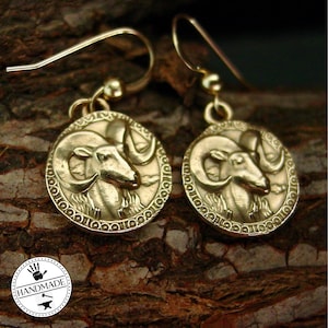May include: Gold-toned earrings with a detailed ram head design. The ram's head is in relief and has a textured surface. The earrings are on a wooden background.