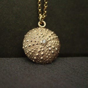 A round domed gold sea urchin shell pendant hanging from a thin gold chain. The charm has a lots of texture and a sparkly faceted white gemstone in its center.
