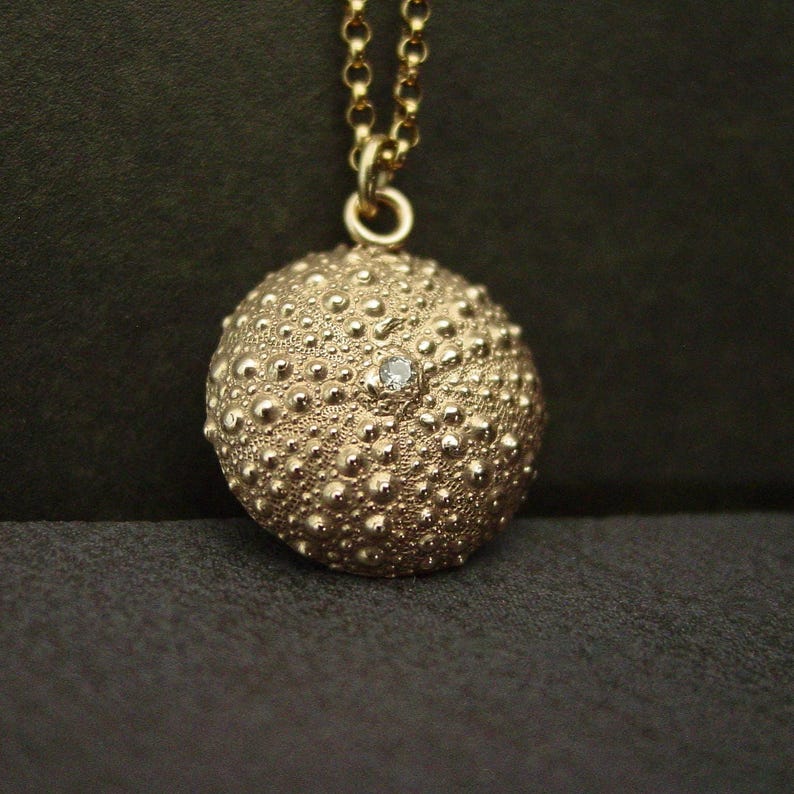A round domed gold sea urchin shell pendant hanging from a thin gold chain. The charm has a lots of texture and a sparkly faceted white gemstone in its center.