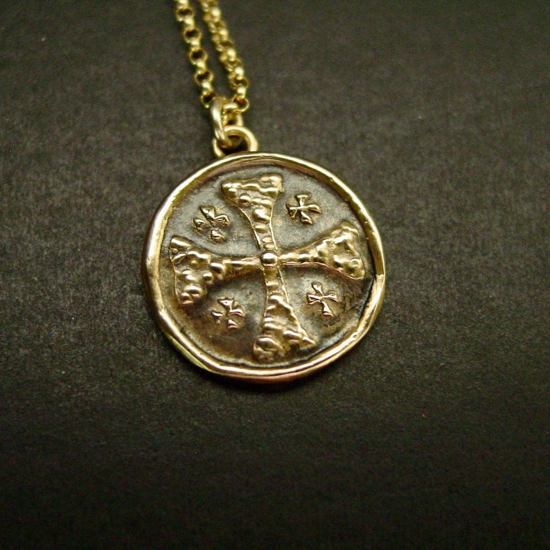 May include: A gold-toned circular pendant necklace featuring a textured cross design in the centre. The pendant is suspended from a matching gold-toned chain. The surface of the pendant has a dark, recessed background with four small cross symbols.