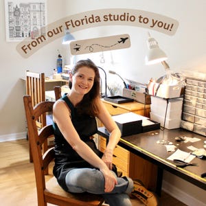 May include: A woman seated in a studio, smiling. The workspace includes a desk with tools, a wooden chair, and a sign that reads "From our Florida studio to you!" with airplane graphics. The room is well-lit.