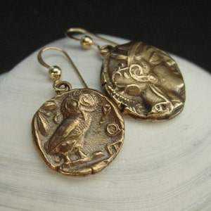 May include: A pair of bronze-colored earrings. Each earring features a circular pendant with a detailed relief design. One pendant depicts an owl, while the other shows a profile of a face. The earrings have gold-colored ear wires.