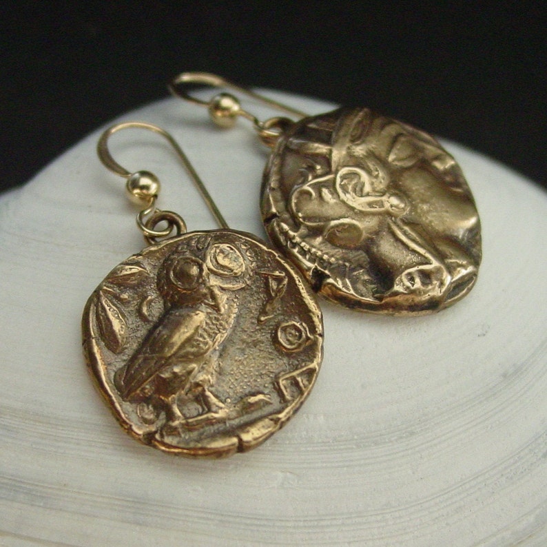 May include: A pair of bronze-colored earrings. Each earring features a circular pendant with a detailed relief design. One pendant depicts an owl, while the other shows a profile of a face. The earrings have gold-colored ear wires.