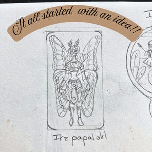 May include: A graphite pencil sketch on paper featuring a central rectangular frame containing a figure with butterfly wings, a skull-like face, and ornate clothing. The text "It all started with an idea!!" appears above, and "Itz papalotl" is written below the figure.
