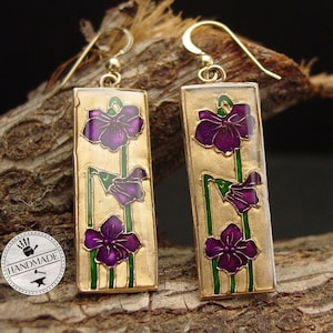 May include: Handmade gold-tone earrings with a rectangular design. Each earring features a purple flower design with green stems and leaves on a gold background. The earrings are hanging on a wooden surface.