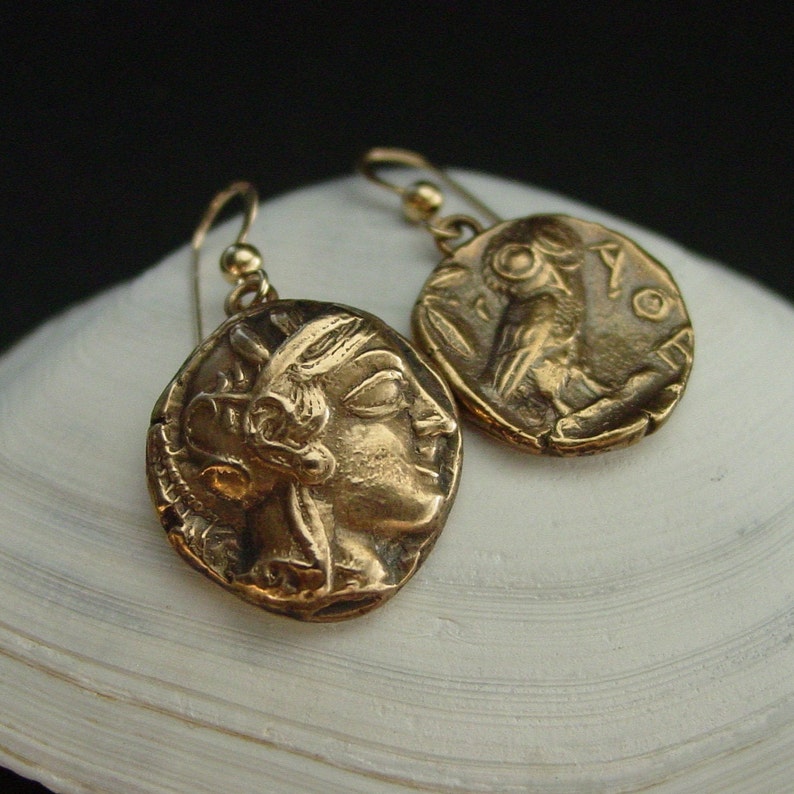 A pair of antiqued bronze coin earrings with uneven edges with cracks from being ancient. On the charms are two different figures, one is the profile of Athena wearing her helmet and on the other charm is the Athenian owl with a palm leaf on the left side and the letters AOE on the right side.