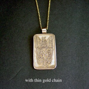 May include: A rectangular gold-toned pendant featuring an engraved illustration of a figure with large butterfly wings, suspended from a thin gold-toned chain. The pendant has a smooth, metallic finish and displays the text with thin gold chain.