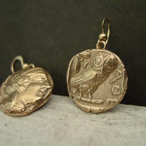 May include: Gold-toned metal earrings. One earring features a profile of a face, the other depicts an owl with Greek lettering. The earrings have a hook closure. The earrings are on a light-colored surface against a dark background.