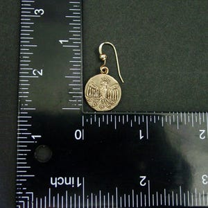 May include: A gold-colored earring with a circular pendant featuring a detailed relief of a bird with outstretched wings. The earring has a hook closure. The pendant is approximately 0.75 inches in diameter.