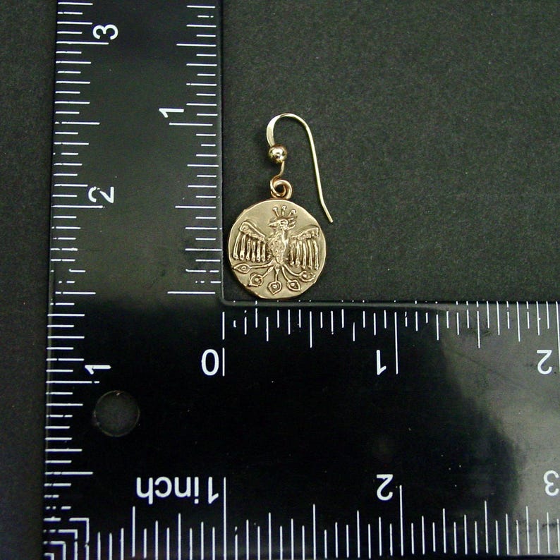 May include: A gold-colored earring with a circular pendant featuring a detailed relief of a bird with outstretched wings. The earring has a hook closure. The pendant is approximately 0.75 inches in diameter.