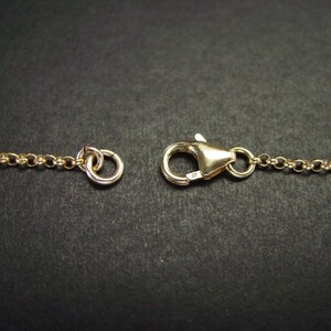 May include: Close-up of a gold-coloured necklace clasp and chain. The clasp is a lobster claw style, and the chain is a delicate link design. The background is a dark, solid colour.