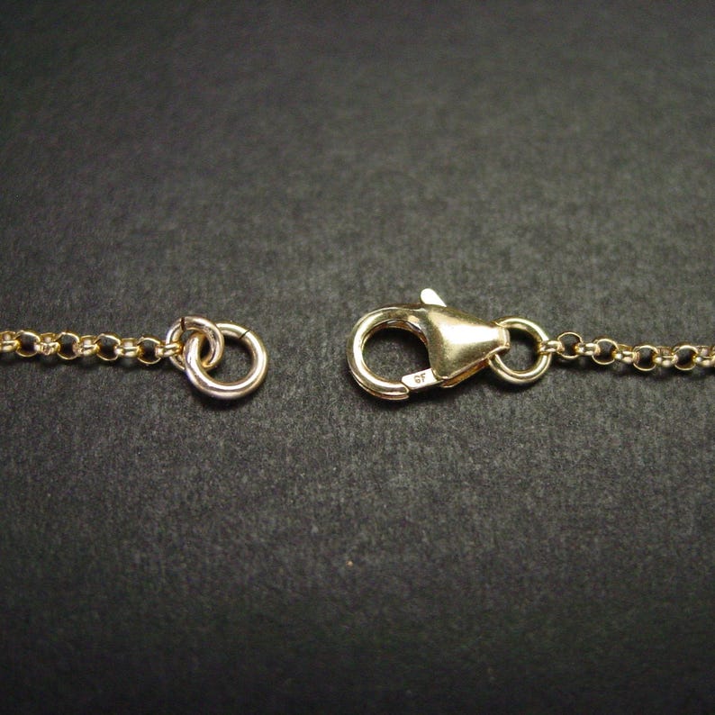 May include: Close-up of a gold-coloured necklace clasp and chain. The clasp is a lobster claw style, and the chain is a delicate link design. The background is a dark, solid colour.