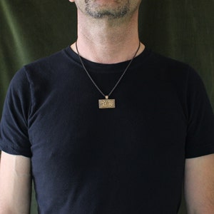 K&ouml;nnte beinhalten: Ein Mann tr&auml;gt ein schwarzes T-Shirt mit einer schwarzen Schnurkette und einem goldenen Anh&auml;nger. Der Anh&auml;nger ist rechteckig und hat ein detailliertes Design.