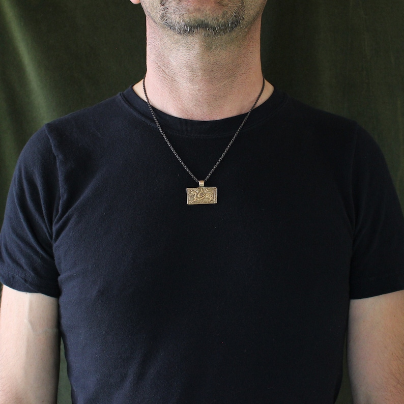 K&ouml;nnte beinhalten: Ein Mann tr&auml;gt ein schwarzes T-Shirt mit einer schwarzen Schnurkette und einem goldenen Anh&auml;nger. Der Anh&auml;nger ist rechteckig und hat ein detailliertes Design.
