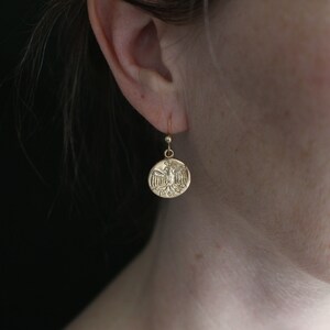 May include: Gold-toned drop earring featuring a circular pendant with an embossed eagle design. The earring hangs from a simple hook. The jewelry is displayed against a dark background, highlighting the intricate details of the pendant.