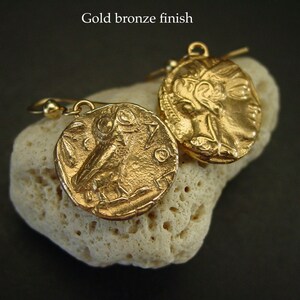 May include: Gold bronze finish earrings. Each earring features a circular pendant with a detailed relief design. One pendant depicts an owl, while the other showcases a profile. The earrings are displayed on a textured, off-white surface.