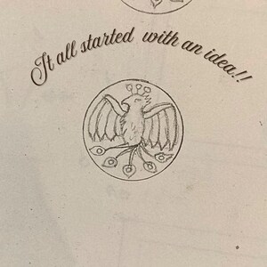 May include: A graphite pencil sketch on off-white paper featuring a circular emblem with a stylized bird, possibly a phoenix, with spread wings and a long tail. Above the circular drawing, the text reads, "It all started with an idea!!"
