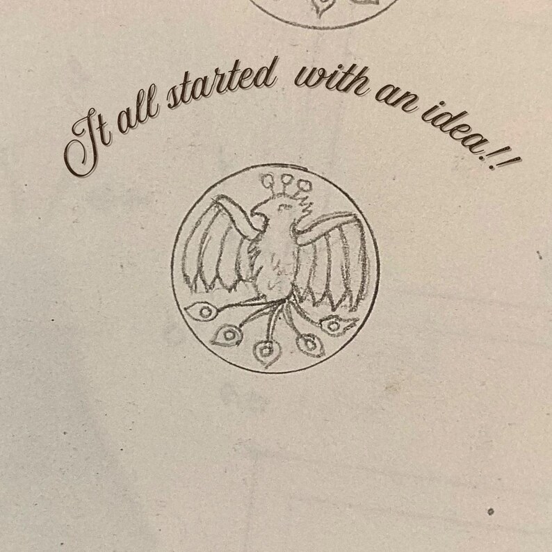 May include: A graphite pencil sketch on off-white paper featuring a circular emblem with a stylized bird, possibly a phoenix, with spread wings and a long tail. Above the circular drawing, the text reads, "It all started with an idea!!"