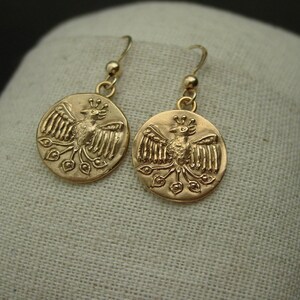 May include: A pair of gold-colored earrings. Each earring features a circular pendant with a detailed relief of a bird with outstretched wings and a crown. The earrings have a simple hook design for pierced ears.