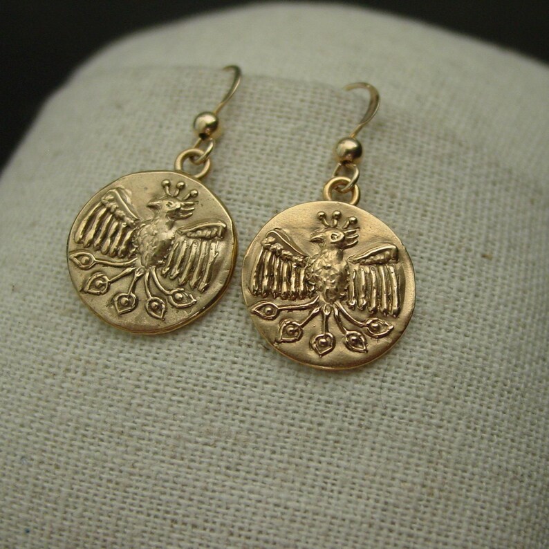 May include: A pair of gold-colored earrings. Each earring features a circular pendant with a detailed relief of a bird with outstretched wings and a crown. The earrings have a simple hook design for pierced ears.
