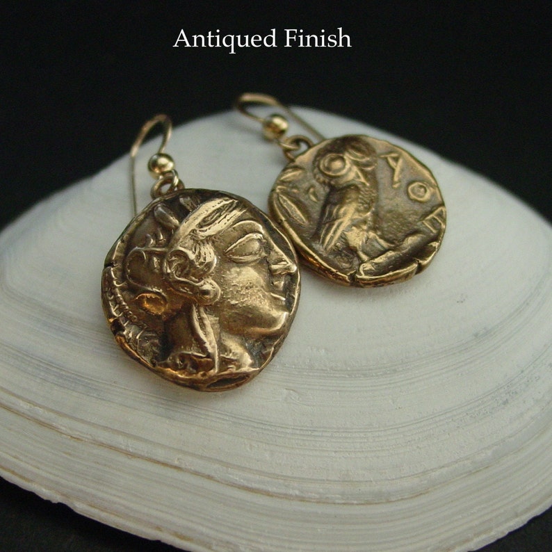 May include: Gold-toned earrings with an antique finish, featuring coin-like pendants. Each pendant displays a detailed relief of a classical figure. The earrings hang from simple ear wires. The text "Antiqued Finish" is visible.